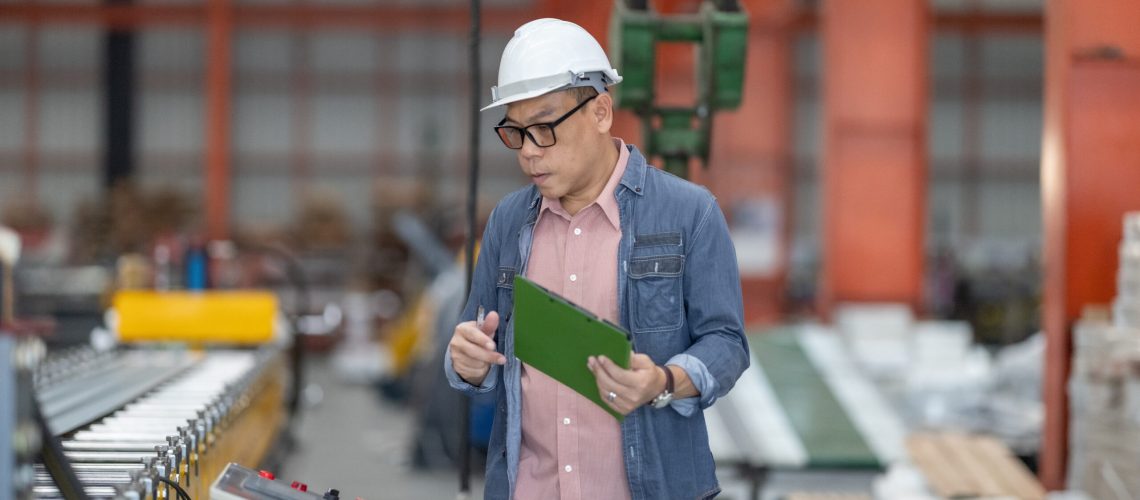 An experienced supervisor meticulously reviews data on a tablet while conducting a quality control check amidst the machinery of a metal sheet factory