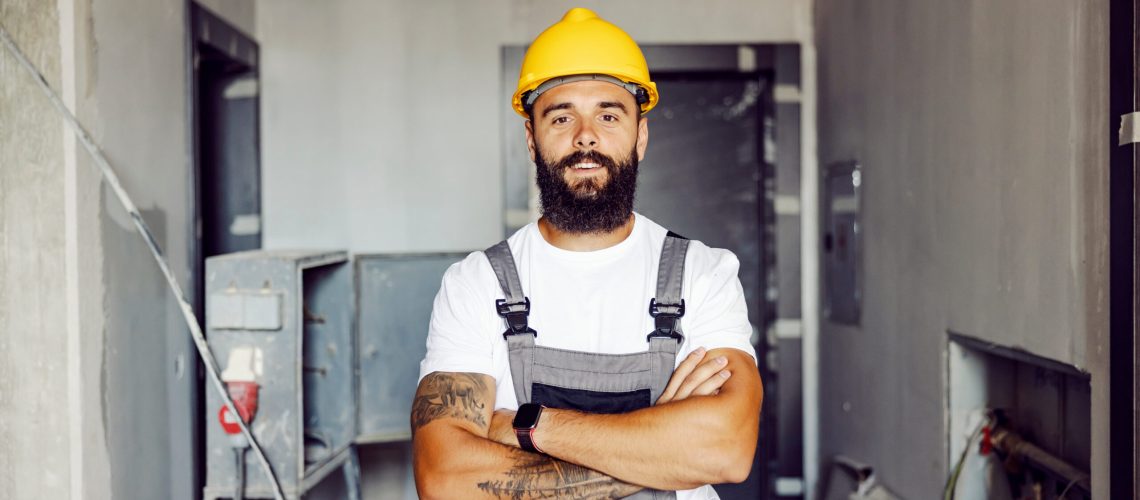 A proud blue collar worker with helmet holding arms crossed at workplace.